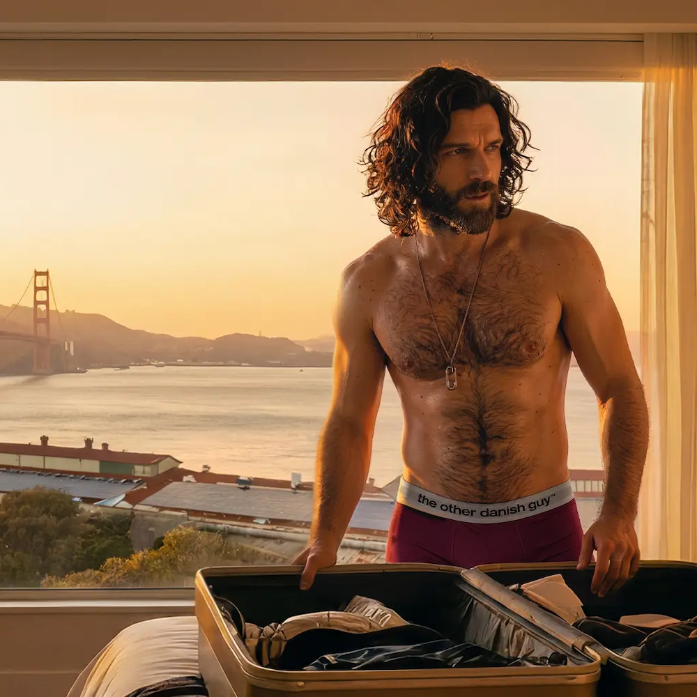 Man wearing royal purple underwear while packing a suitcase at sunrise, with the Golden Gate Bridge visible in the background.
